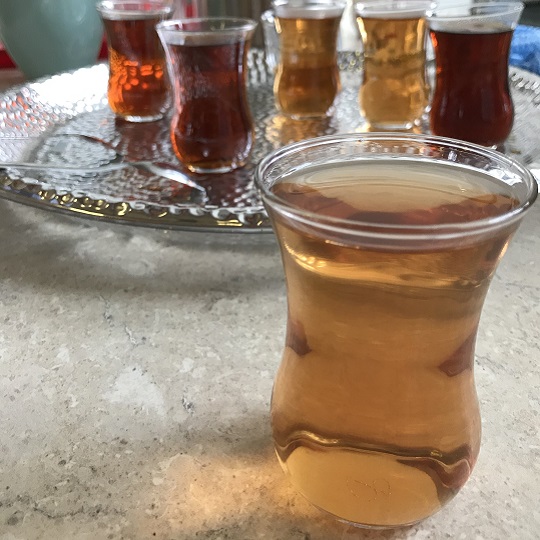 Photo of little Turkish clear tulip tea cups made of glass, one glass with clear light tea colour sits on a large round silver trey, a number of small Turkish glass cups sit in the background, some with a darker, deeper red colour tea